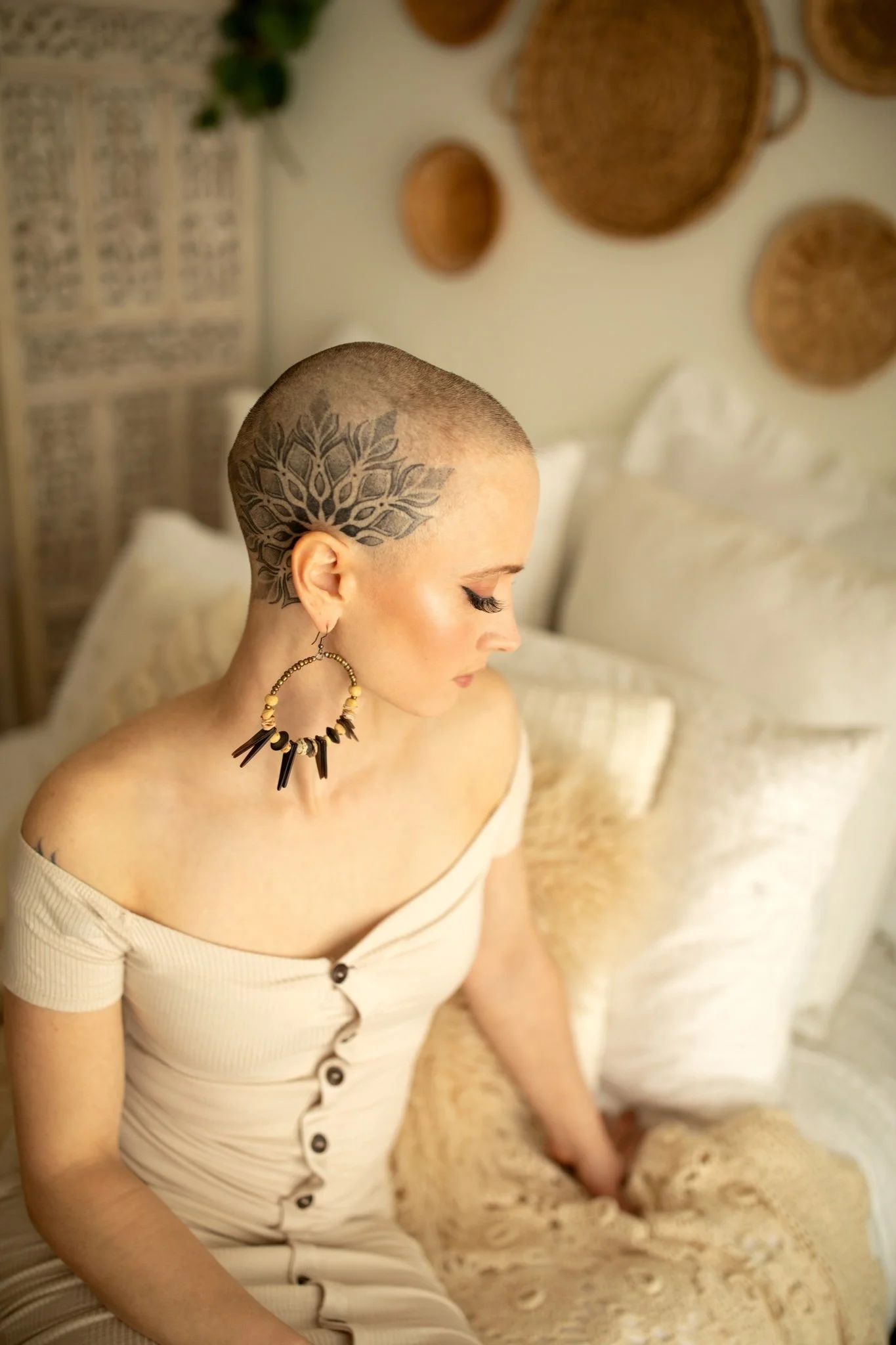 A woman with a shaved head and a large floral tattoo on her scalp, sitting on a bed with cream-colored bedding, wearing a beige, off-the-shoulder, button-down dress and a beaded black and beige necklace.