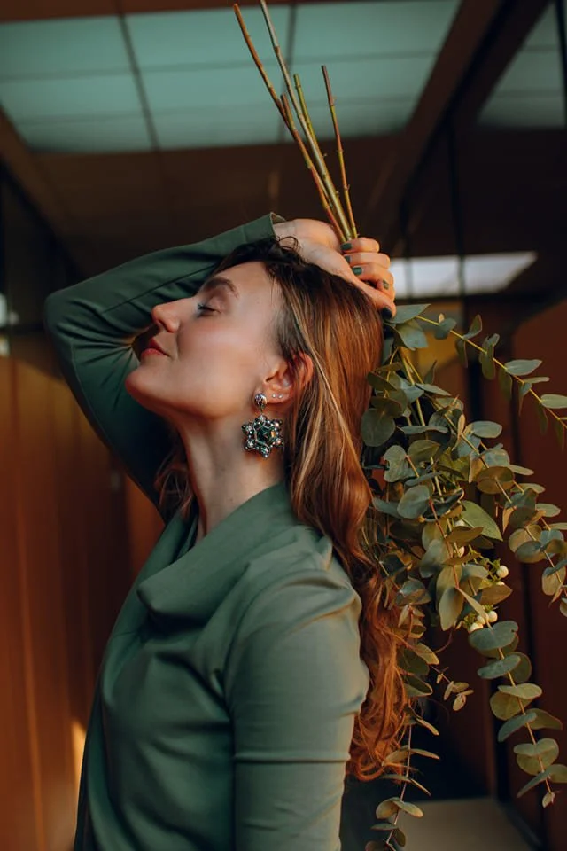 A woman with red hair and earrings, wearing a green top, is posing with her eyes closed and her head tilted back, holding a bundle of sticks and greenery behind her head.