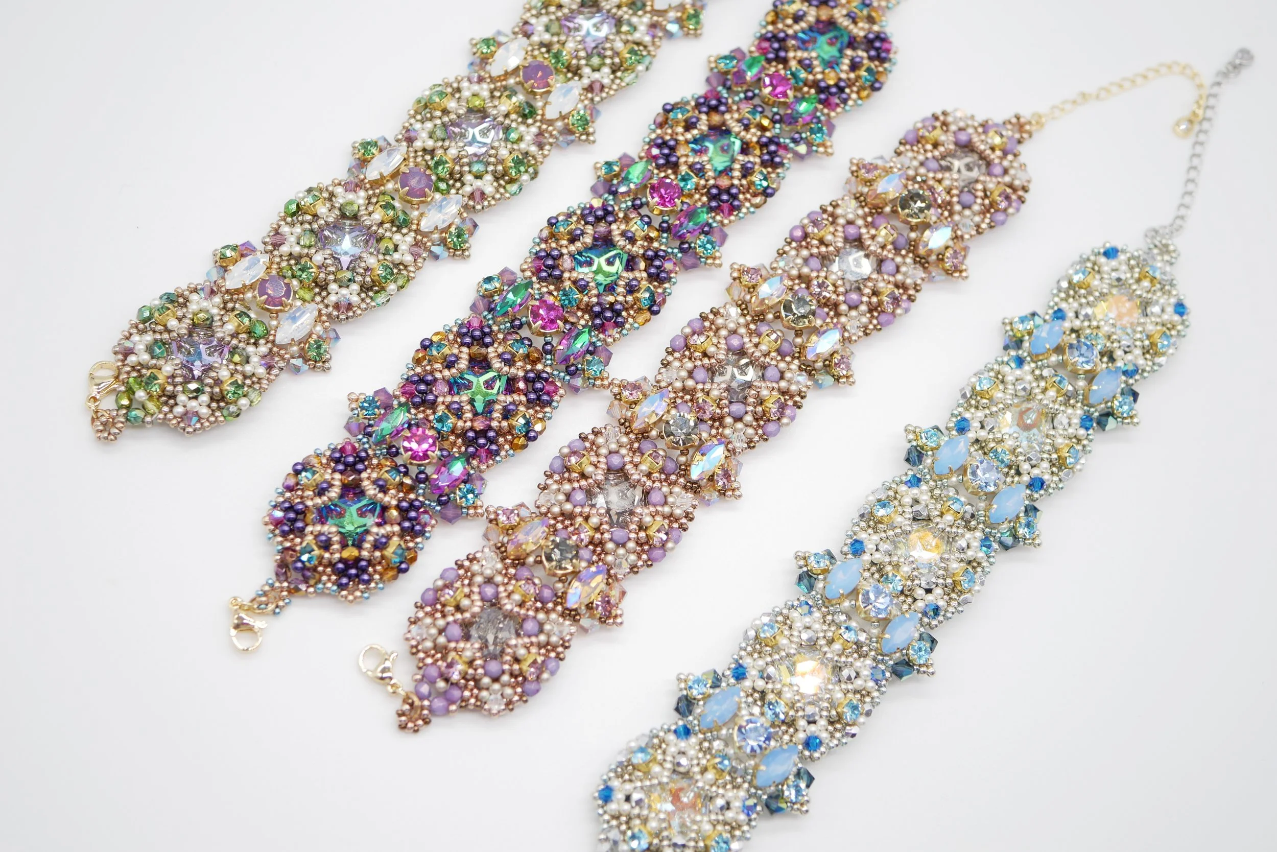 Four colorful, ornate beaded necklaces with rhinestones, pearls, and intricate designs laid out on a white surface.