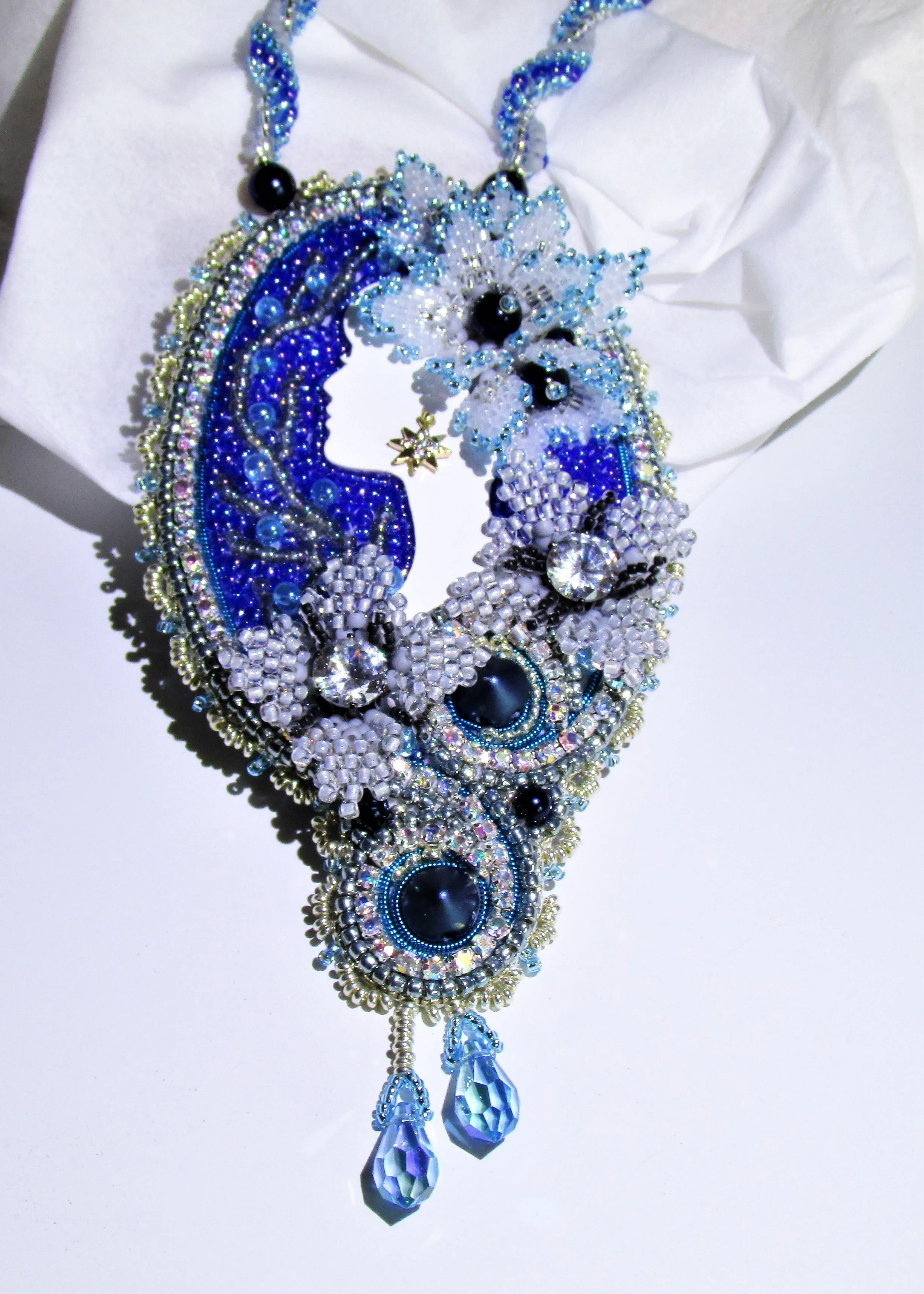 A large, intricate jewelry necklace with blue and white beads, black accents, and crystal embellishments, featuring a moon and star design and dangling crystal pendants.