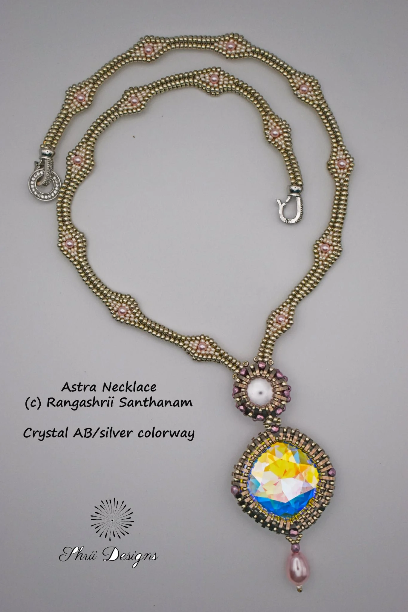 A beaded necklace with a circular pendant featuring a large multicolored crystal, accented with pink and silver beads, attached to a chain of alternating pink and gold beads; the necklace has a hook clasp and is labeled 'Astra Necklace' by Rangashrii