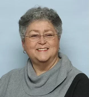 Smiling elderly woman with gray hair, glasses, and earrings, wearing a gray scarf and black top against a light blue background.