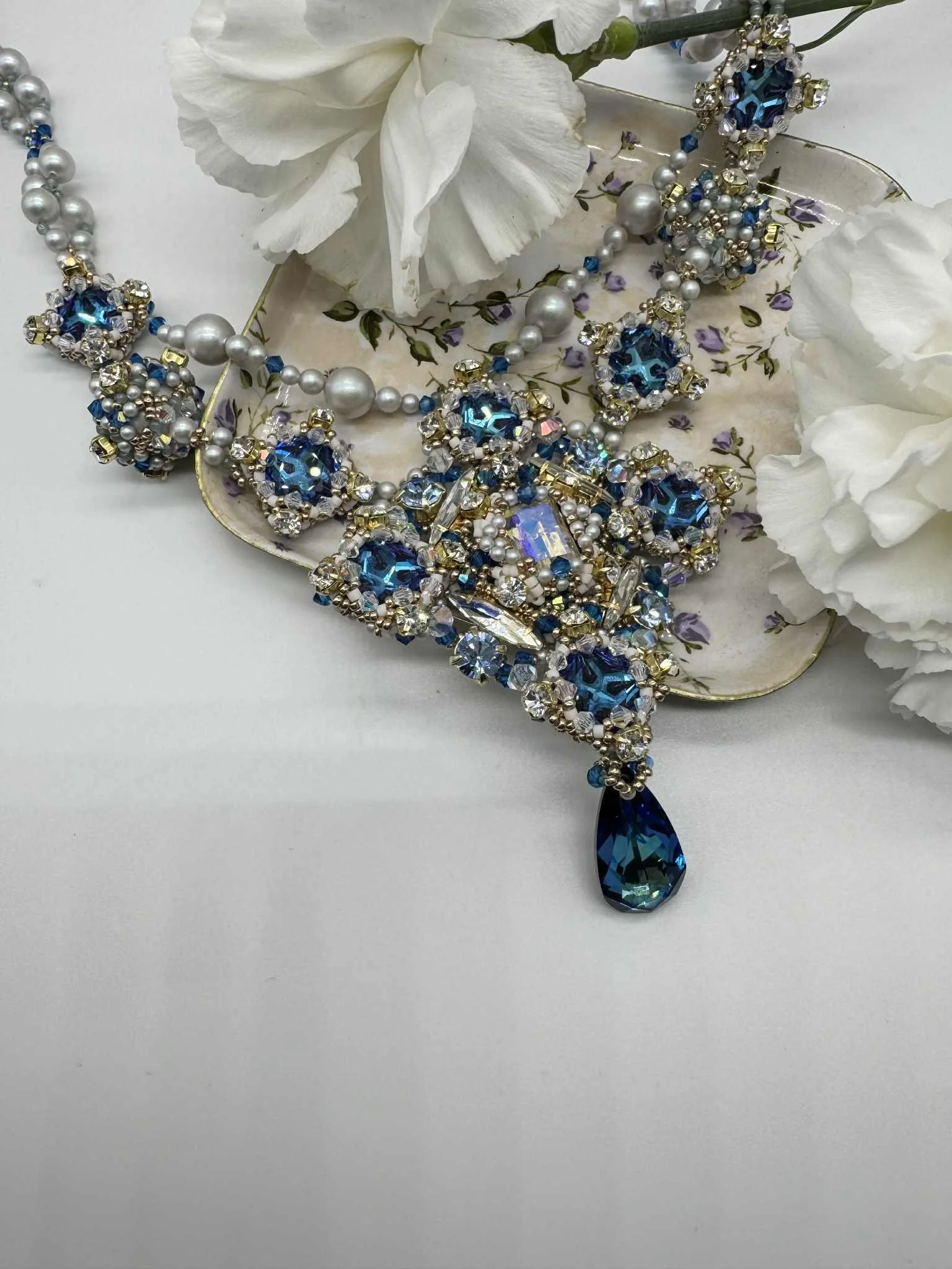 A jewelry necklace with blue, white, and gold beads, adorned with large blue and iridescent gemstones, resting on a decorative plate with floral patterns, surrounded by white artificial flowers.
