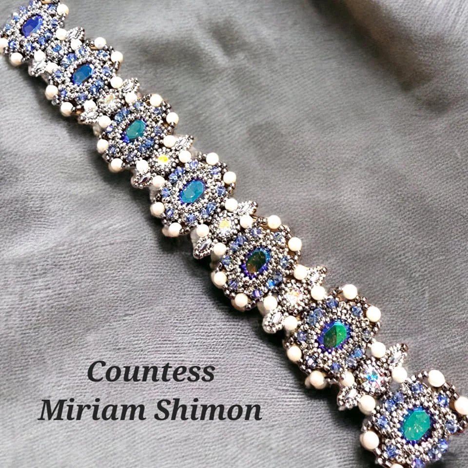 A detailed silver bracelet with blue, green, and white gemstones, and pearl accents, placed on gray fabric. Text overlay reads "Countess Miriam Shimon."