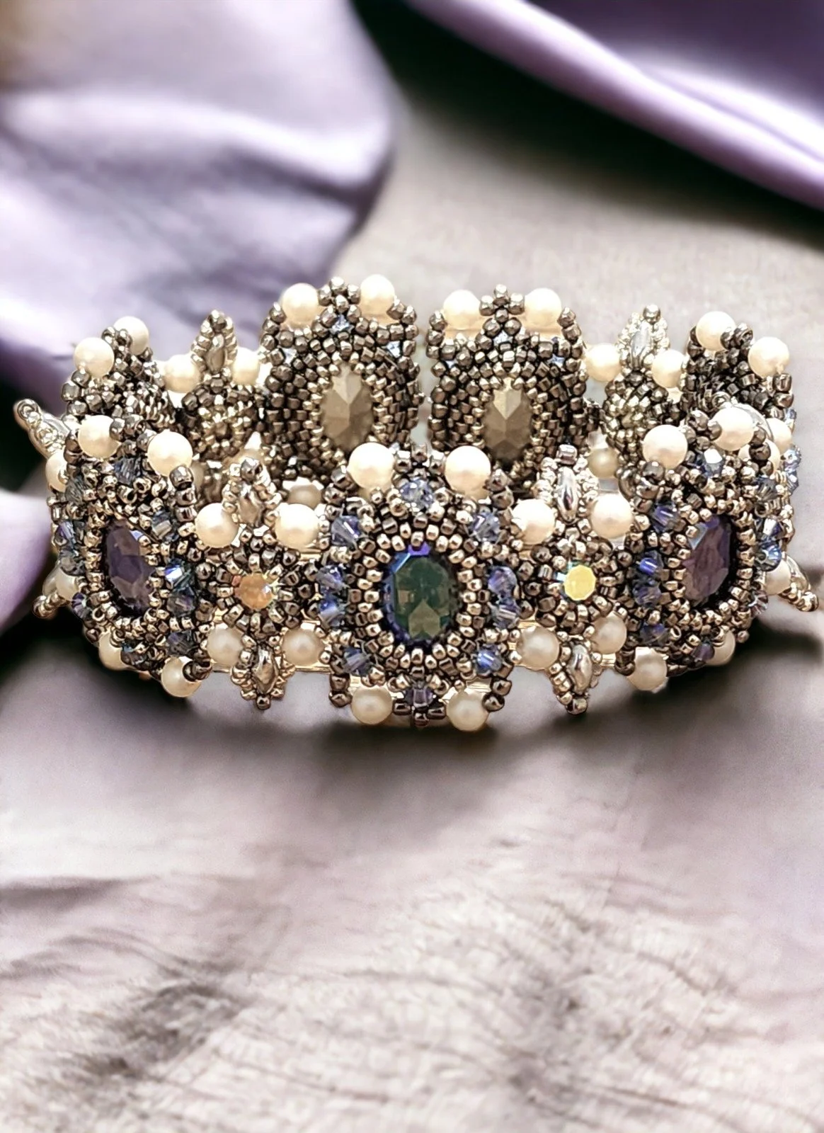 An ornate tiara with pearls, rhinestones, and large oval and round gemstones in purple, green, and amber colors, resting on a soft fabric surface.