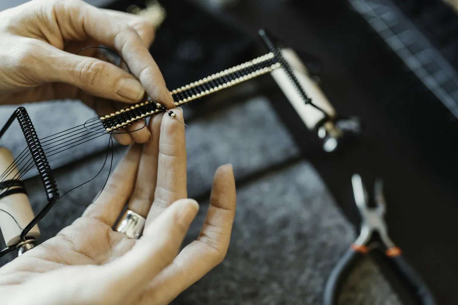 Close-up of hands threading small beads onto a string as part of jewelry making, with tools and a beading loom visible.
