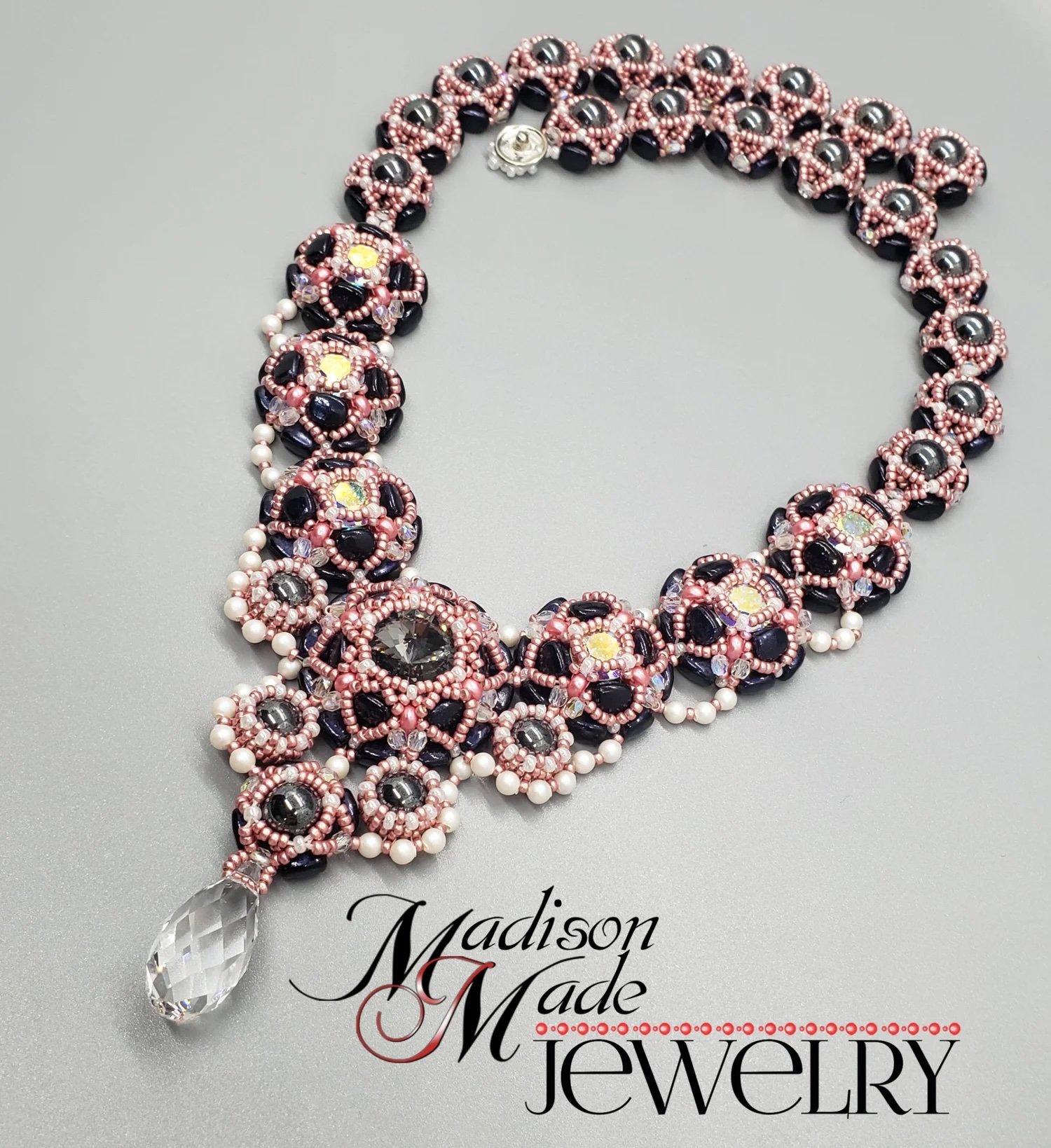 A beaded necklace with pink, black, and pearl beads, featuring a large drop-shaped crystal pendant and a flower-shaped clasp, placed on a gray surface with the words "Madison Made Jewelry" printed below.