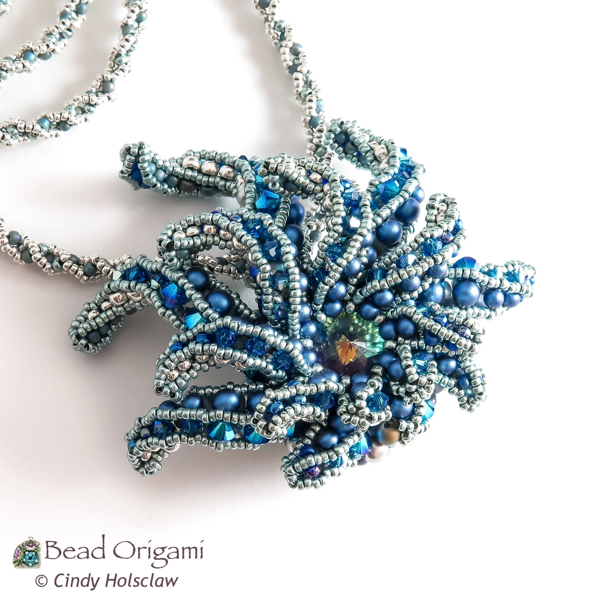 A close-up photo of a handmade bead origami necklace with blue metallic beads, grey beads, and clear beads with a blue tint, designed in a twisted and intertwined pattern.
