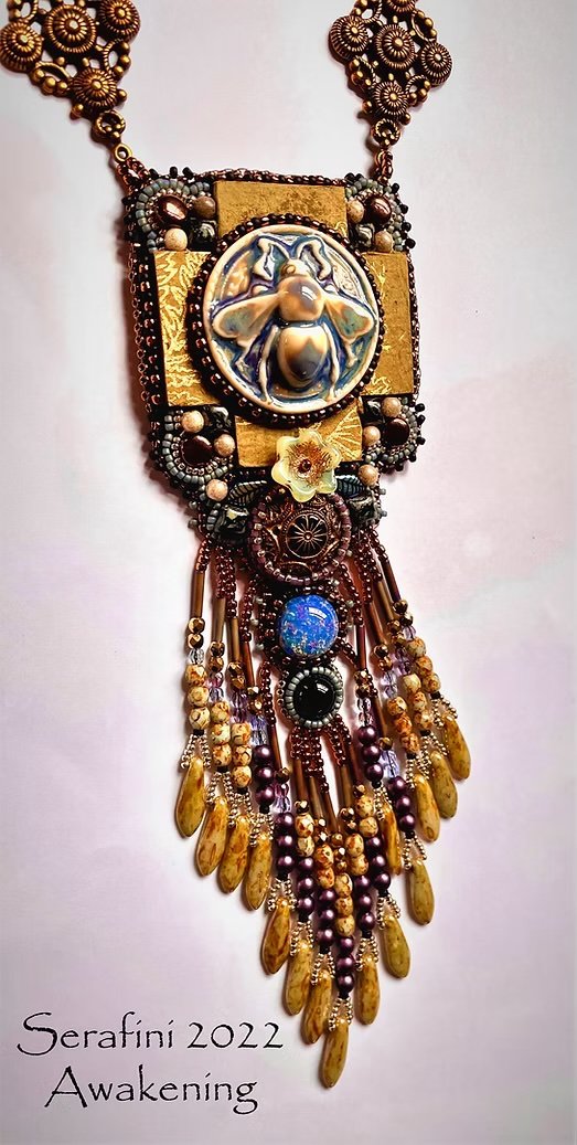 A large, ornate necklace with a bee motif at the center, surrounded by various beads, charms, and dangling elements in earthy tones. The necklace has gold and brown accents and intricate detailing. The bottom left corner has text reading 'Serafini 20