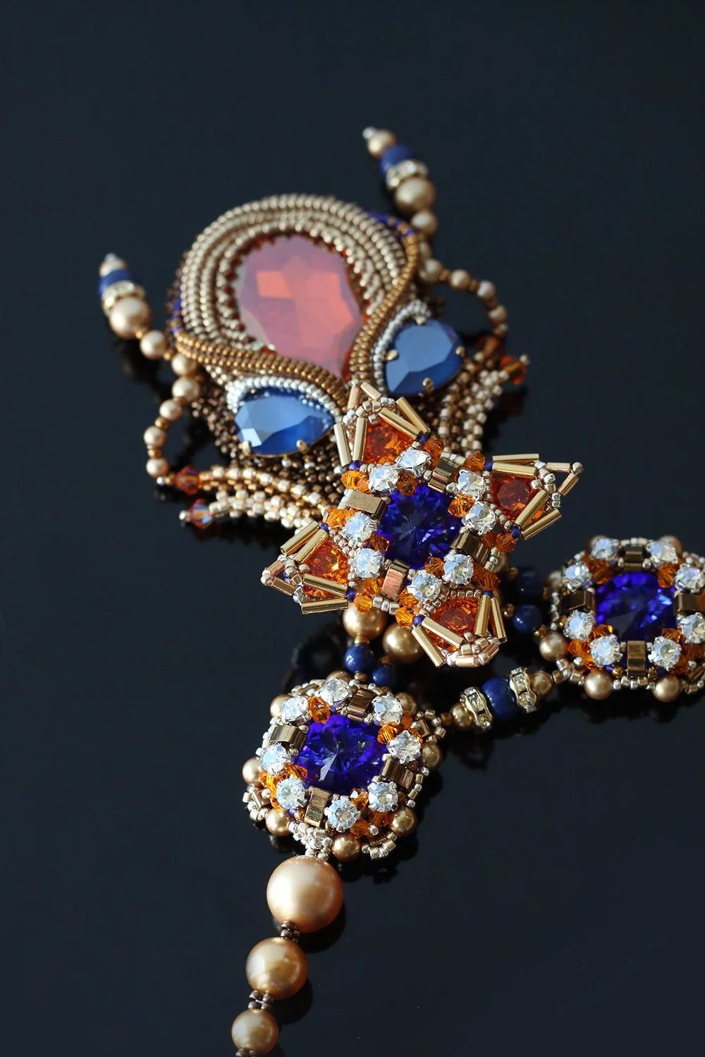 Close-up of a decorative necklace with large pink and blue gemstones, smaller yellow and clear stones, and pearl accents on a black background.