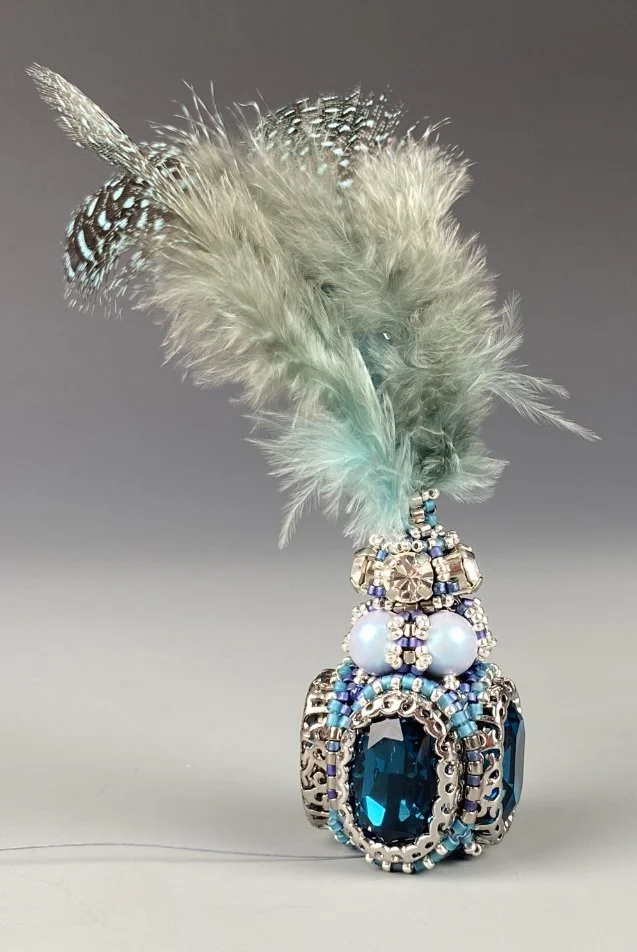 A decorative item with silver, blue, and white beads, large blue gemstones, two white pearls, intricate metalwork, and a cluster of light grey and white feathers at the top.