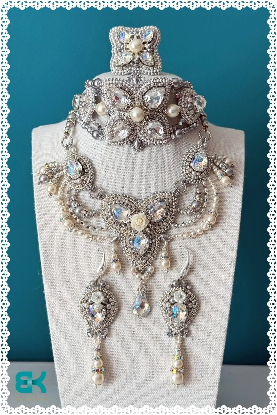 A set of ornate silver jewelry, including a large statement necklace, a matching bracelet, and a tiara or headpiece, all decorated with clear rhinestones, pearls, and floral motifs, displayed on a white fabric bust against a teal background.