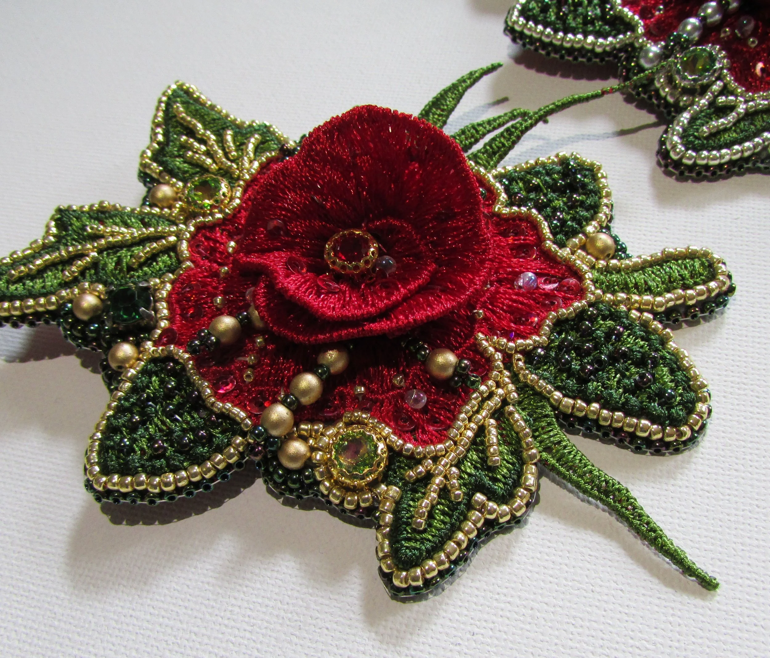 Close-up of a decorative embroidered flower with red petals, green leaves, gold beads, and small shiny embellishments, on a white fabric background.