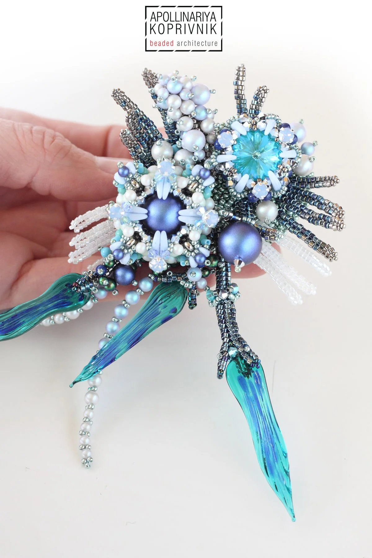Close-up of a beaded brooch designed to resemble a dragonfly, with intricate details including pearl and crystal beads for the body and wings, and two elongated, translucent blue beads for the tail.