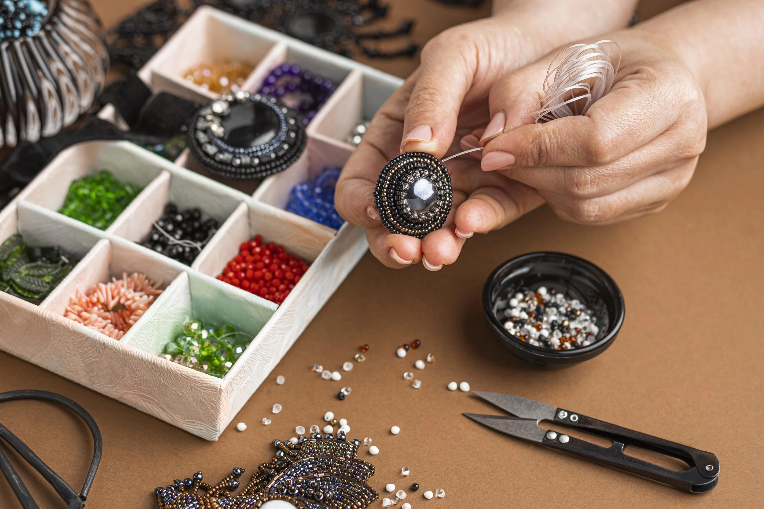 Close-up of hands working on beading jewelry with various beads and jewelry-making tools on a table.