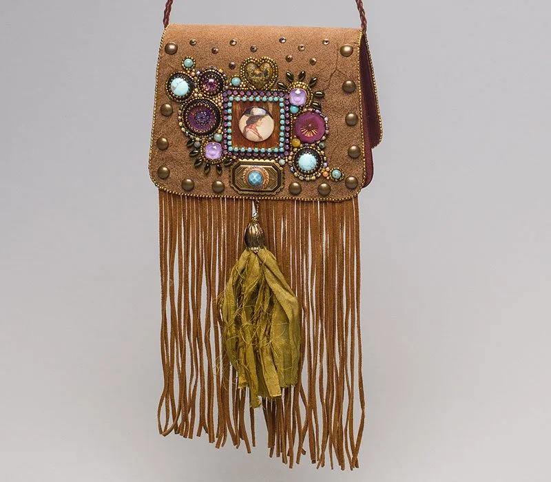 A handbag decorated with various colorful jewelry and beads, featuring a small portrait in the center, with long fringe tassels hanging from the bottom.
