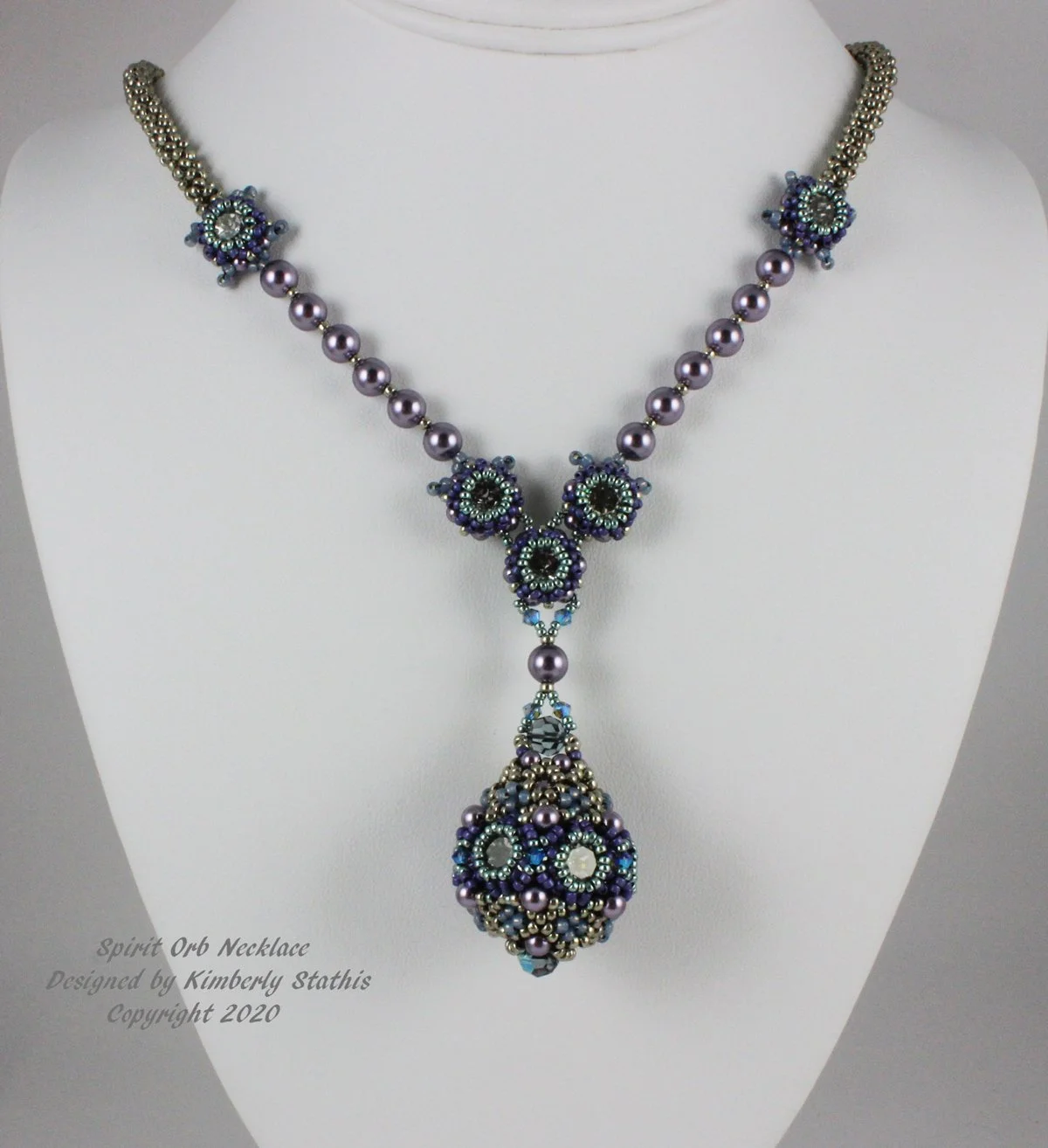 A beaded necklace with purple, blue, and silver beads, featuring a large pendant with intricate beadwork and a pendant drop. The jewelry is displayed on a white bust. Text on the image reads 'Spirit Orb Necklace Designed by Kimberly Stathis Copyright