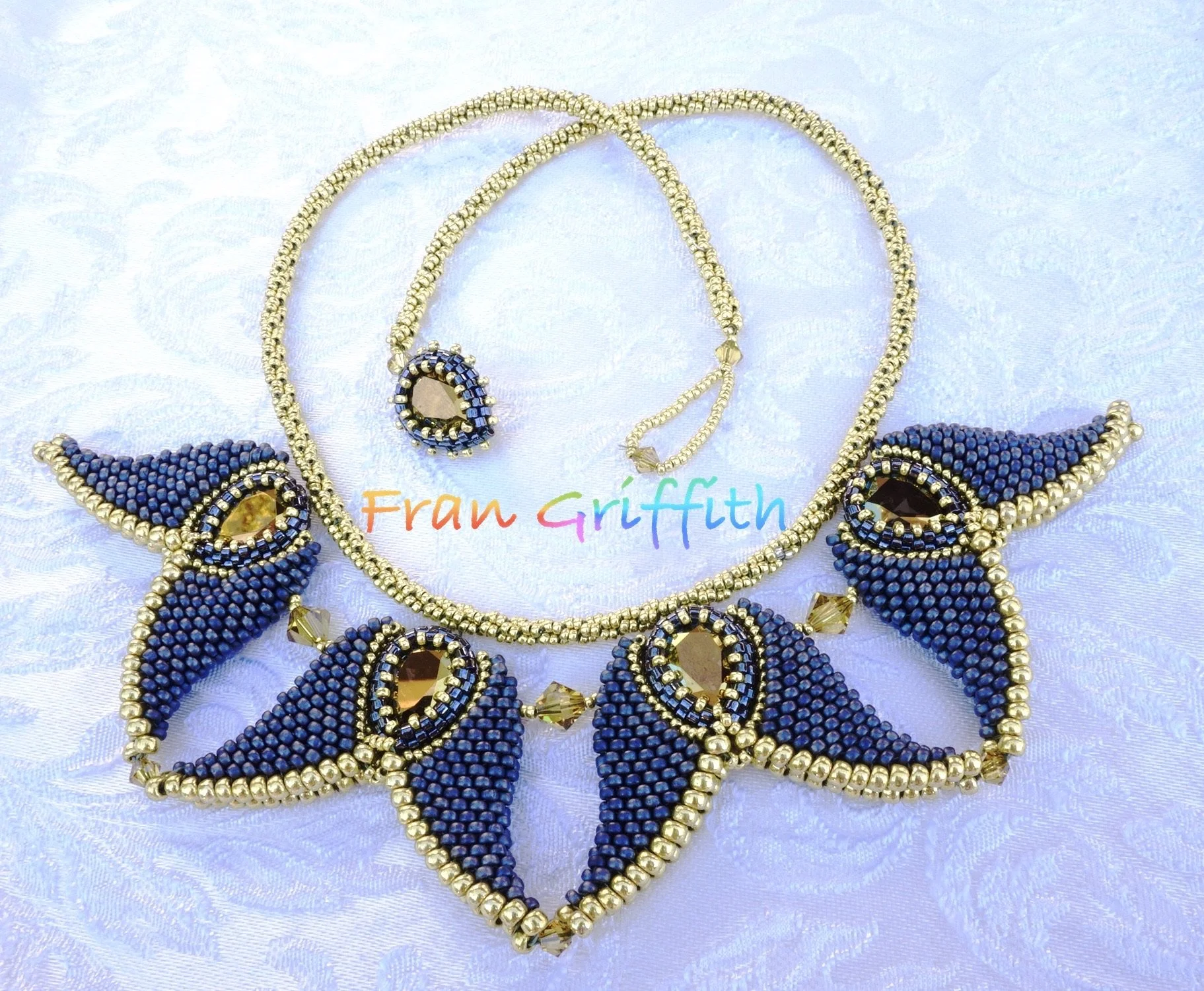 Gold and blue beaded jewelry set including a necklace, bracelet, earrings, and ring with sapphire and gold accents, displayed on light fabric with a 'Fran Griffith' logo.