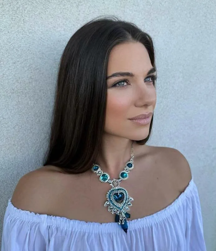 A woman with long brown hair and blue eyes wearing a white off-the-shoulder top and a large necklace with turquoise and blue stones, standing against a plain wall.