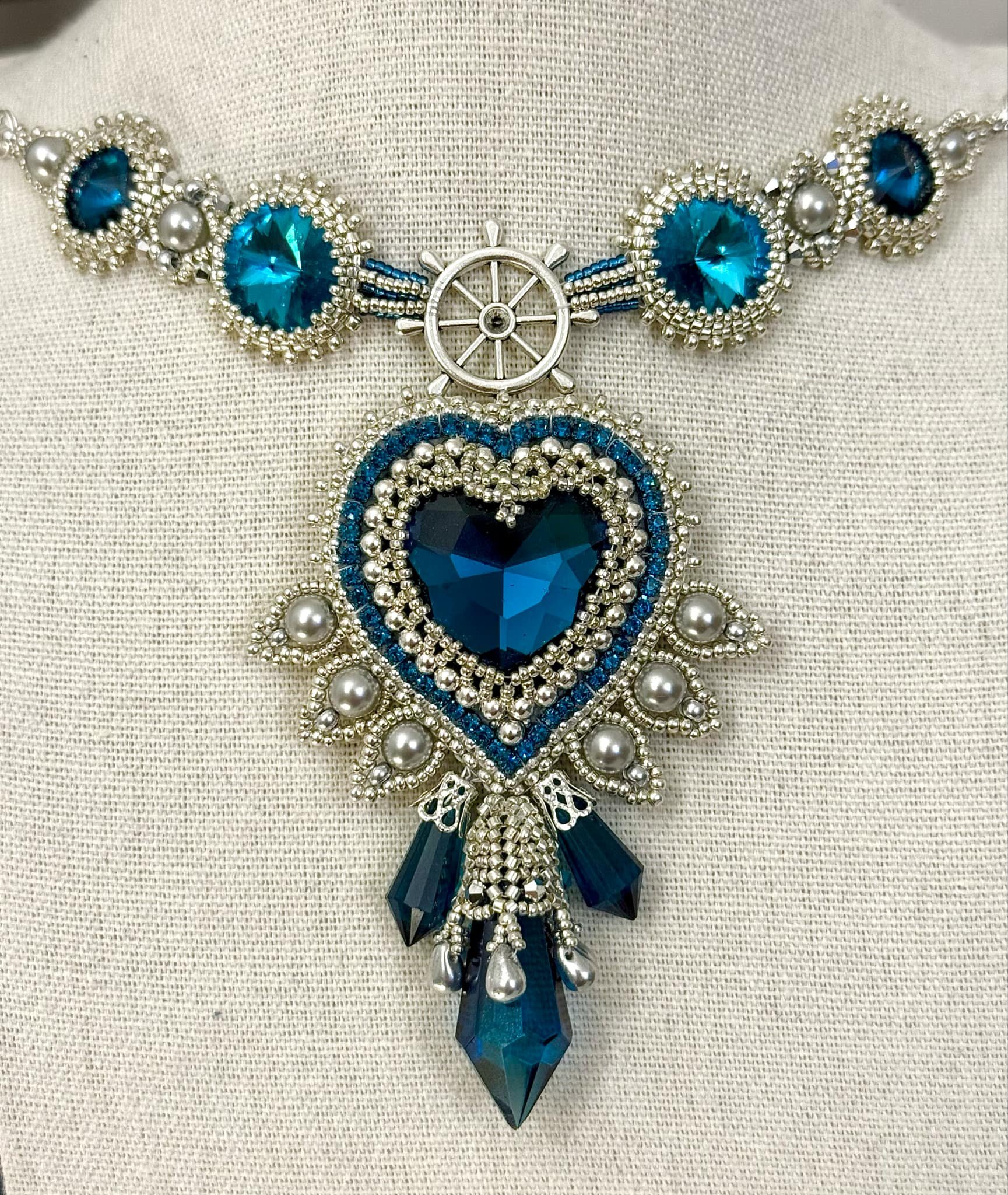 Close-up of an ornate jewelry necklace with blue, silver, and pearl elements on a textured light-colored fabric background.