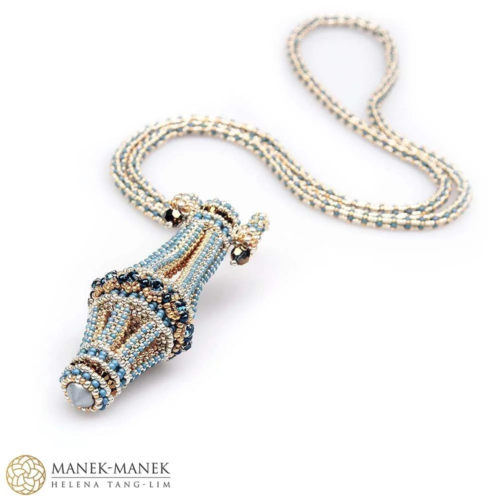 Beaded necklace with a pendant resembling a seashell, made of light blue, gold, and black beads, displayed on a white background. The necklace is from MANEK-MANEK by Helena Tang-Lim.