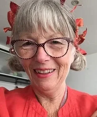 A smiling elderly woman with gray hair, wearing glasses with a red and black polka dot frame, and a red shirt, in a festive setting with red decorations in her hair.