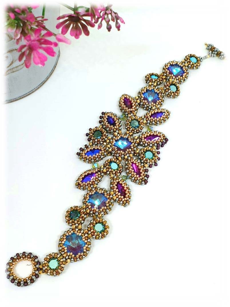 A colorful beaded bracelet with large iridescent and purple stones, adorned with small gold beads, laid on a white surface with a pink flower in the background.