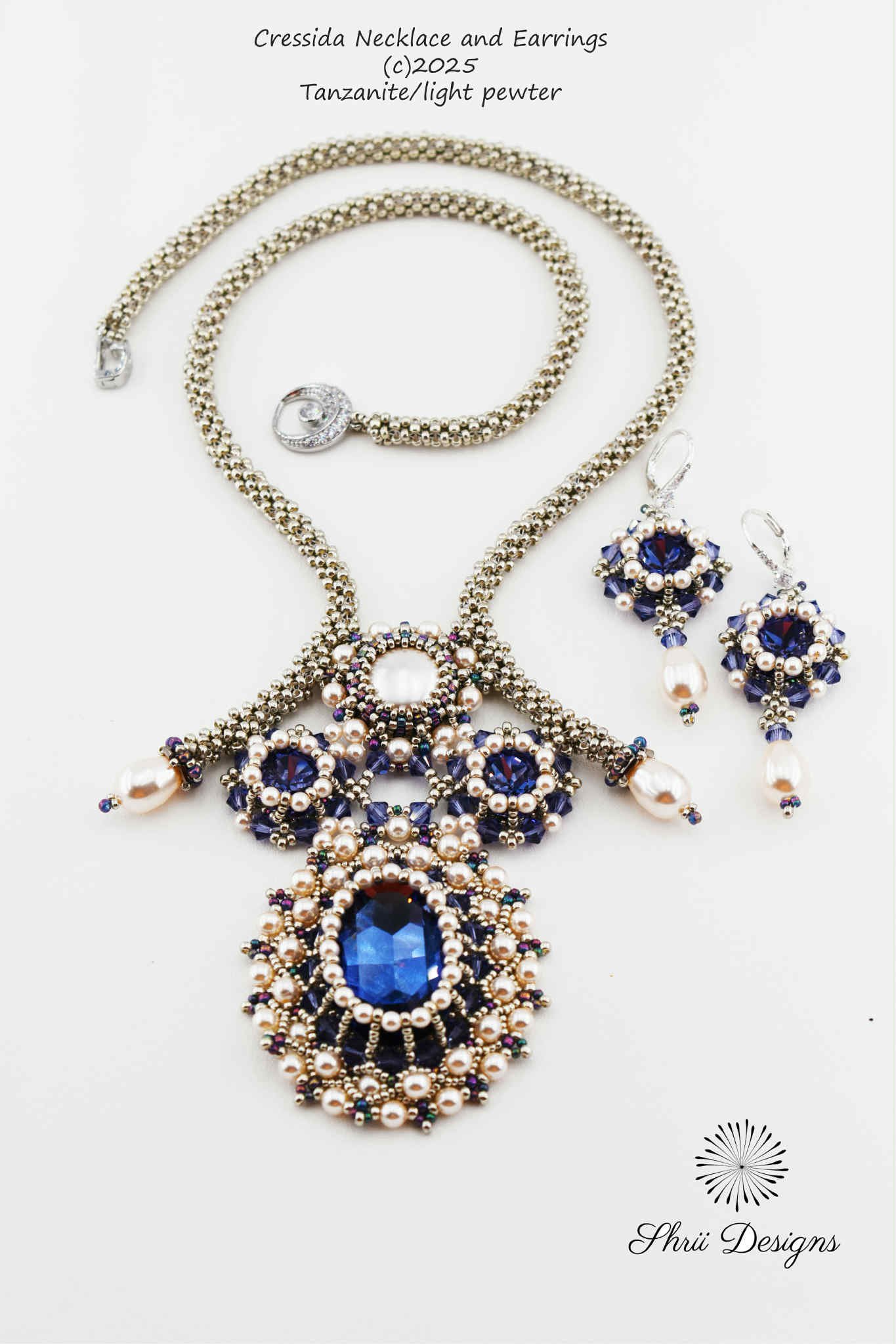 Photo of a jewelry set consisting of a necklace and matching earrings with large blue gemstones, pearls, and intricate beadwork, labeled as Cressida Necklace and Earrings by Shrii Designs, made of tanzanite and light pewter, copyrighted in 2025.