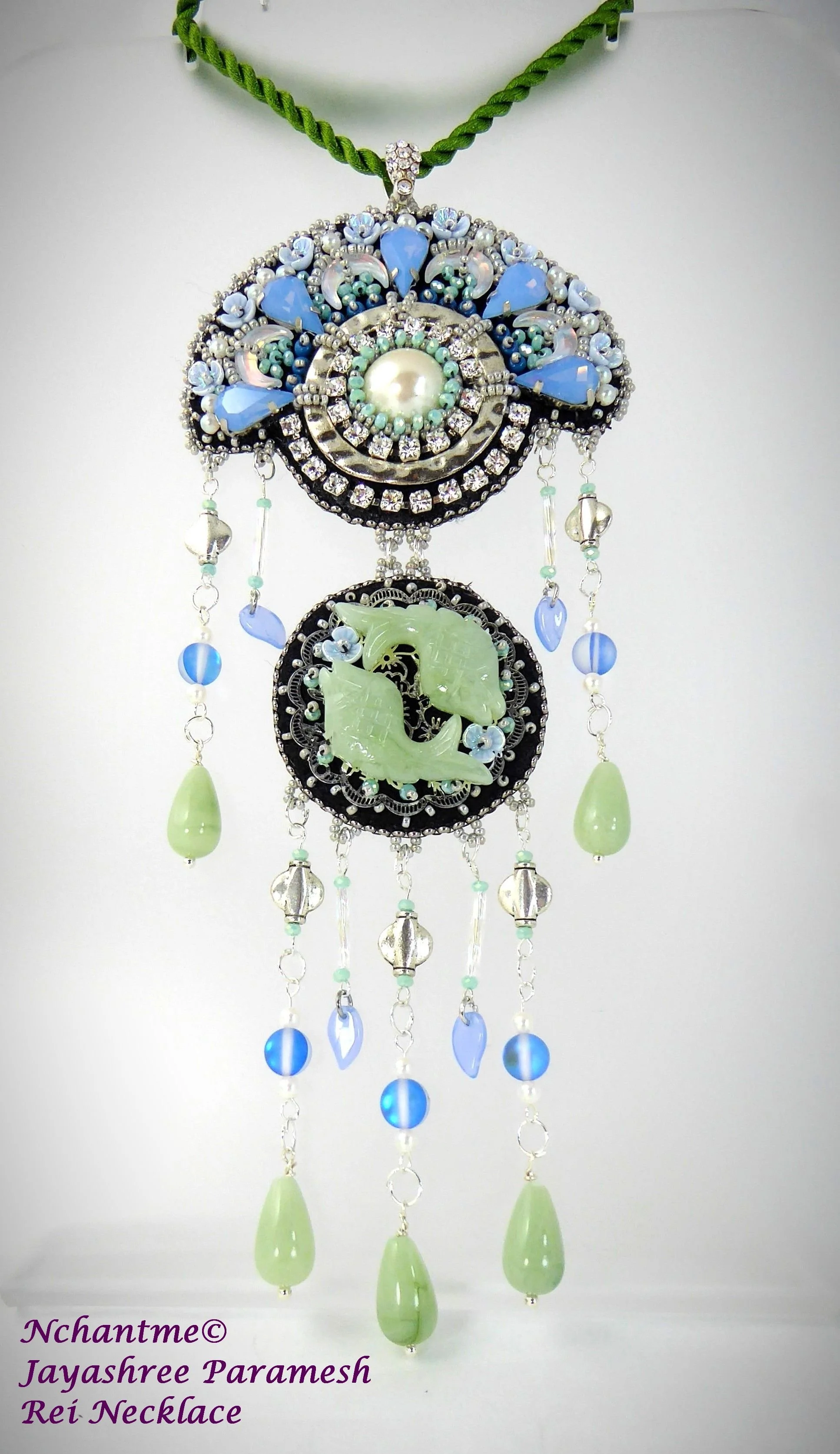 A necklace featuring a large ornate pendant with blue, white, and green beads, rhinestones, and a central pearl, with hanging green and blue teardrop-shaped beads, against a white background, with purple text indicating it is a Jayashree Paramesh Rei
