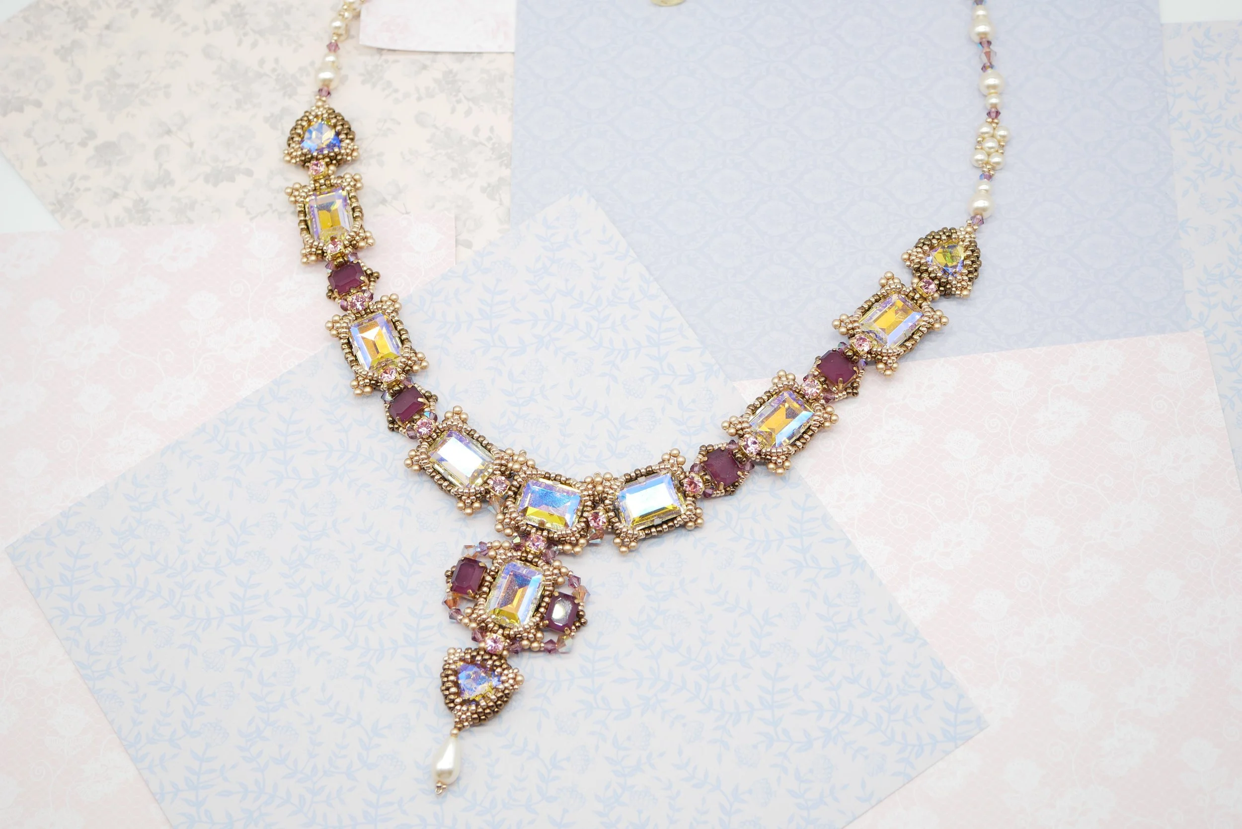 A colorful jeweled necklace with large rectangular and oval stones, gold beads, pink accents, and a dangling pearl at the bottom, laid on patterned pastel papers.