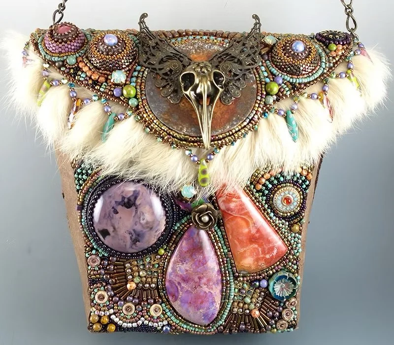 A decorative jewelry box adorned with beads, stones, feathers, and a metal animal skull with wings on top.