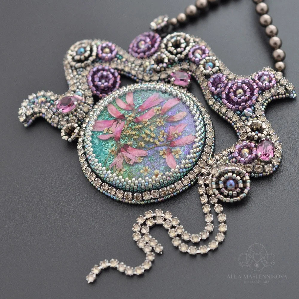 A detailed, ornate necklace with a bejeweled crescent and floral-themed pendant, featuring pink and purple stones, small pearls, and intricate beadwork.