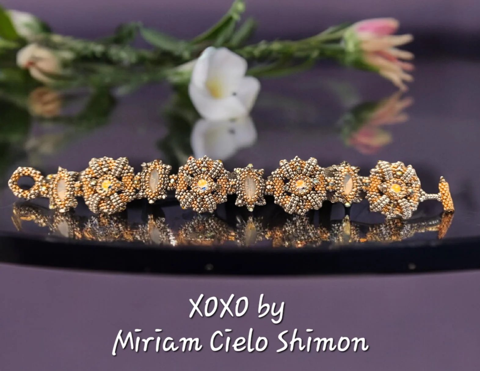 A beaded bracelet with intricate floral designs, placed on a reflective black surface with a blurred white flower and green stem in the background.
