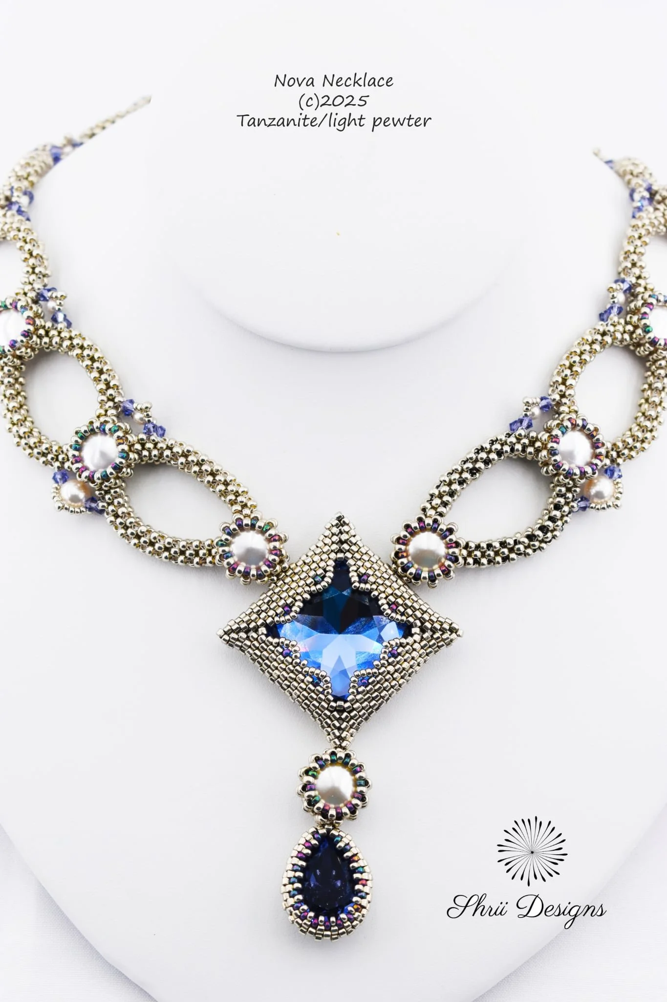 A silver-toned necklace with a large deep blue teardrop-shaped gemstone pendant surrounded by small multicolored beads, displayed on a white bust. The necklace features intricate beaded flower motifs and a star-shaped element with a smaller gemstone 