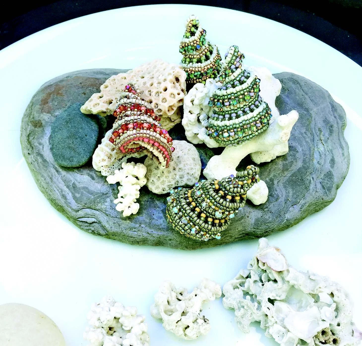 Colorful beaded jewelry displayed on coral and rocks, with additional coral pieces on a white plate.