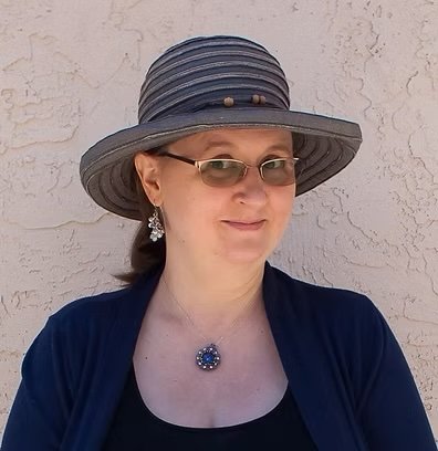 A woman wearing a wide-brimmed striped hat, sunglasses, earrings, and a necklace, standing against a textured beige wall.