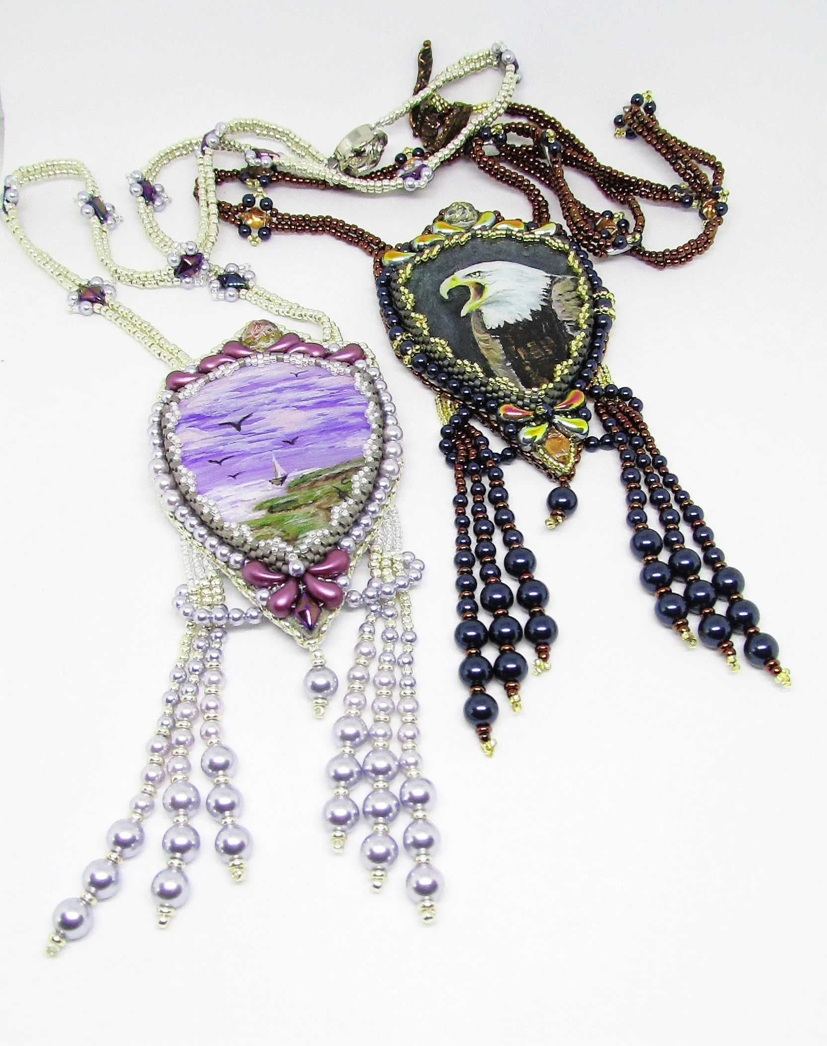 Assorted beaded necklaces, including one with a painted image of an eagle, another with a landscape scene, and others with bead patterns, arranged on a white surface.