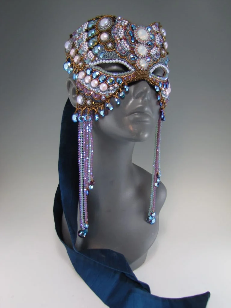 A gray mannequin head wearing an ornate, bejeweled masquerade mask decorated with pearls, gemstones, and beads, with long beaded tassels hanging from the sides, and a dark blue ribbon or fabric draped over the shoulder.