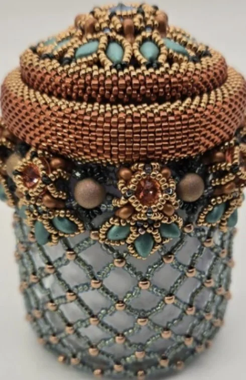 An ornate beaded jewelry box or container with intricate beadwork in shades of copper, teal, black, and gold, featuring floral and geometric patterns.