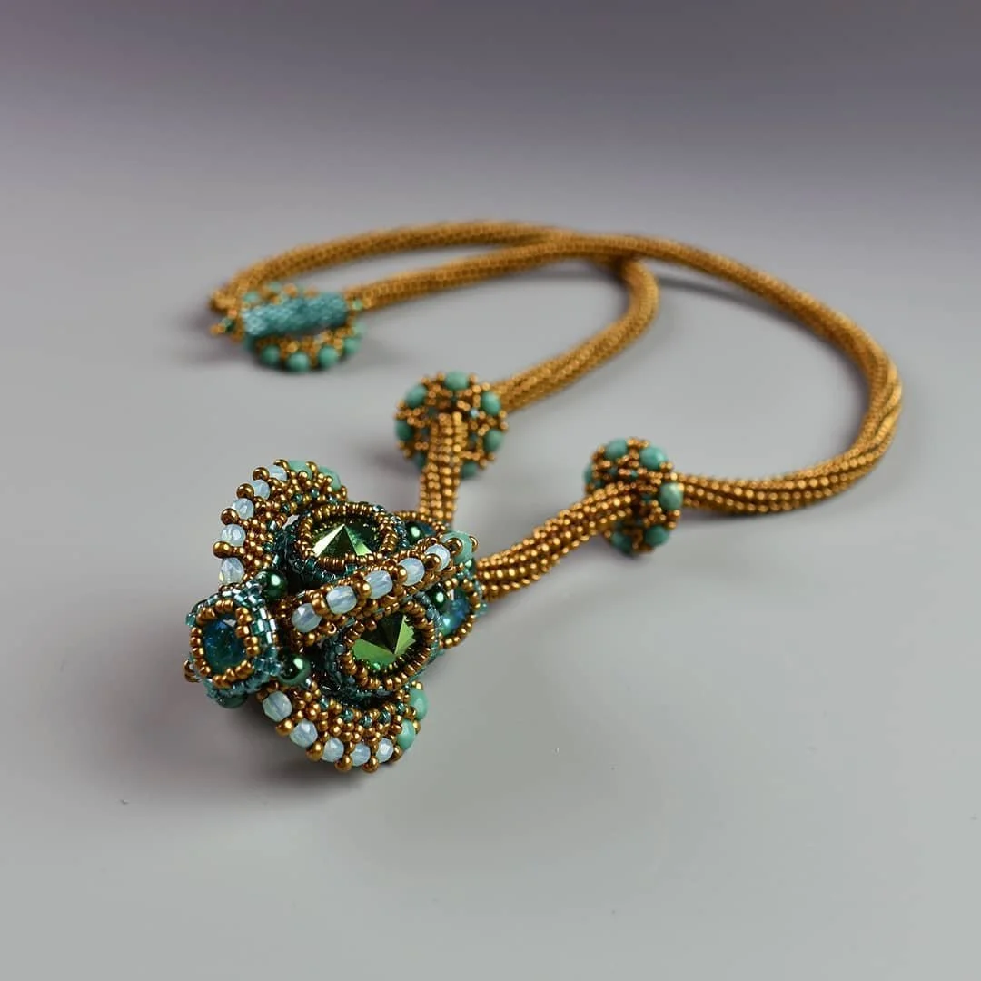 Gold necklace with turquoise and white beads, featuring a large pendant with intricate beadwork and green accents, laid on a gray surface.