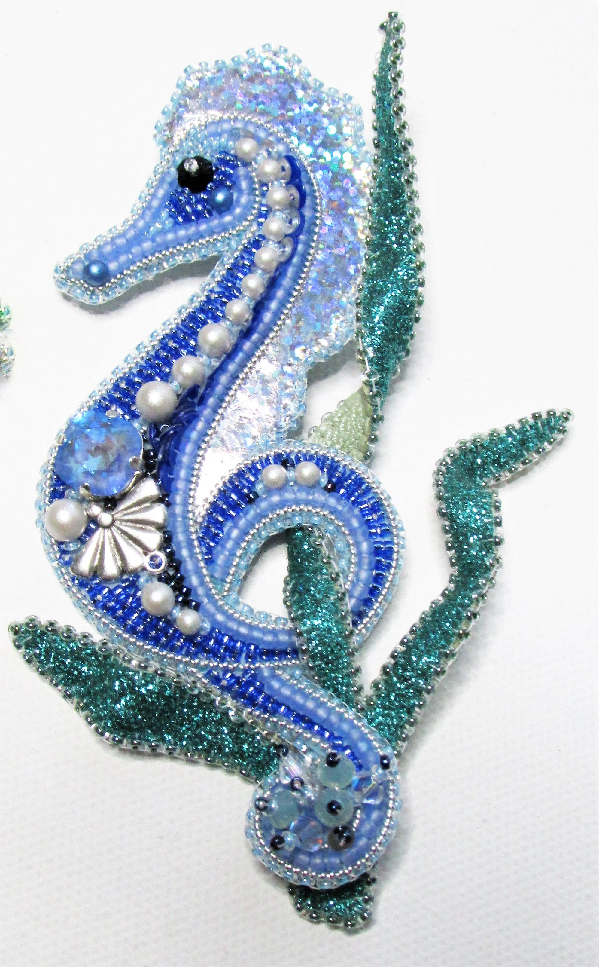 A beaded seahorse brooch with blue, green, white, and black beads, pearls, and rhinestones on a white background.