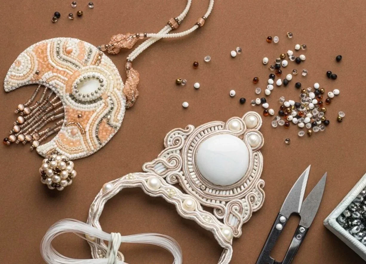 Two intricate necklaces with beads and pearls, a scattered mix of colorful beads, a pair of small scissors, and a box of loose beads on a brown background.