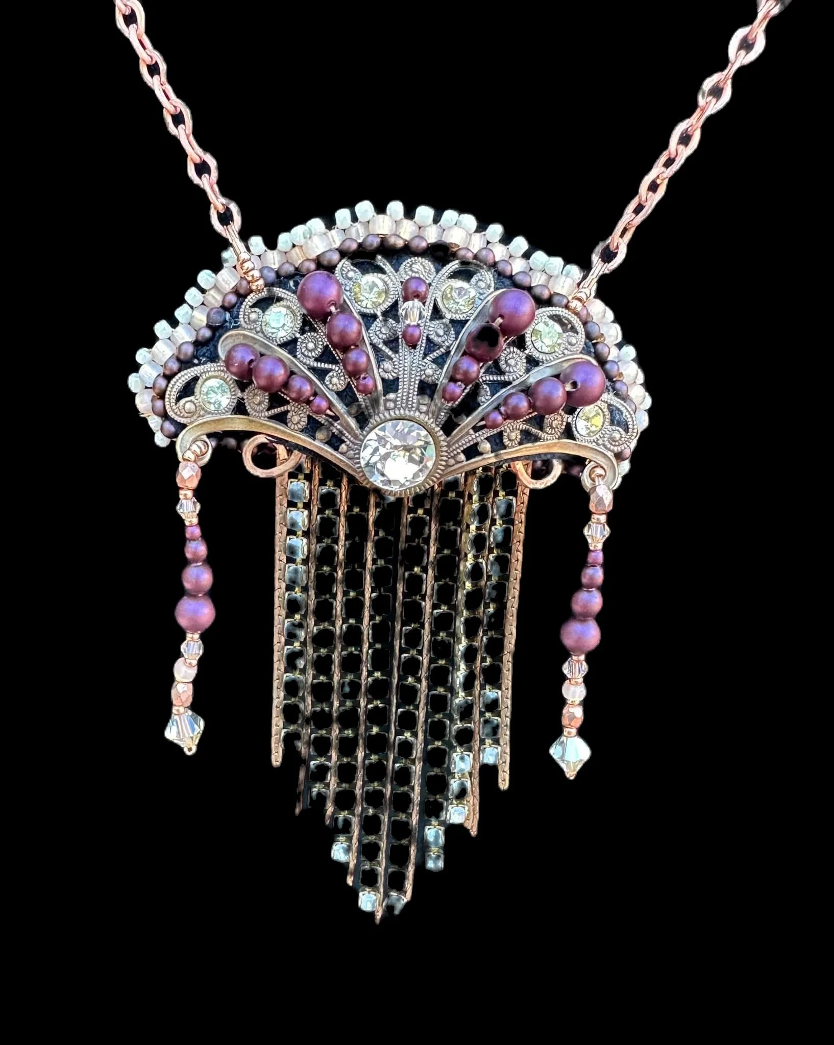 A close-up of an ornate, vintage-style necklace with purple beads, clear rhinestones, and a large central diamond-shaped rhinestone set against a black background.