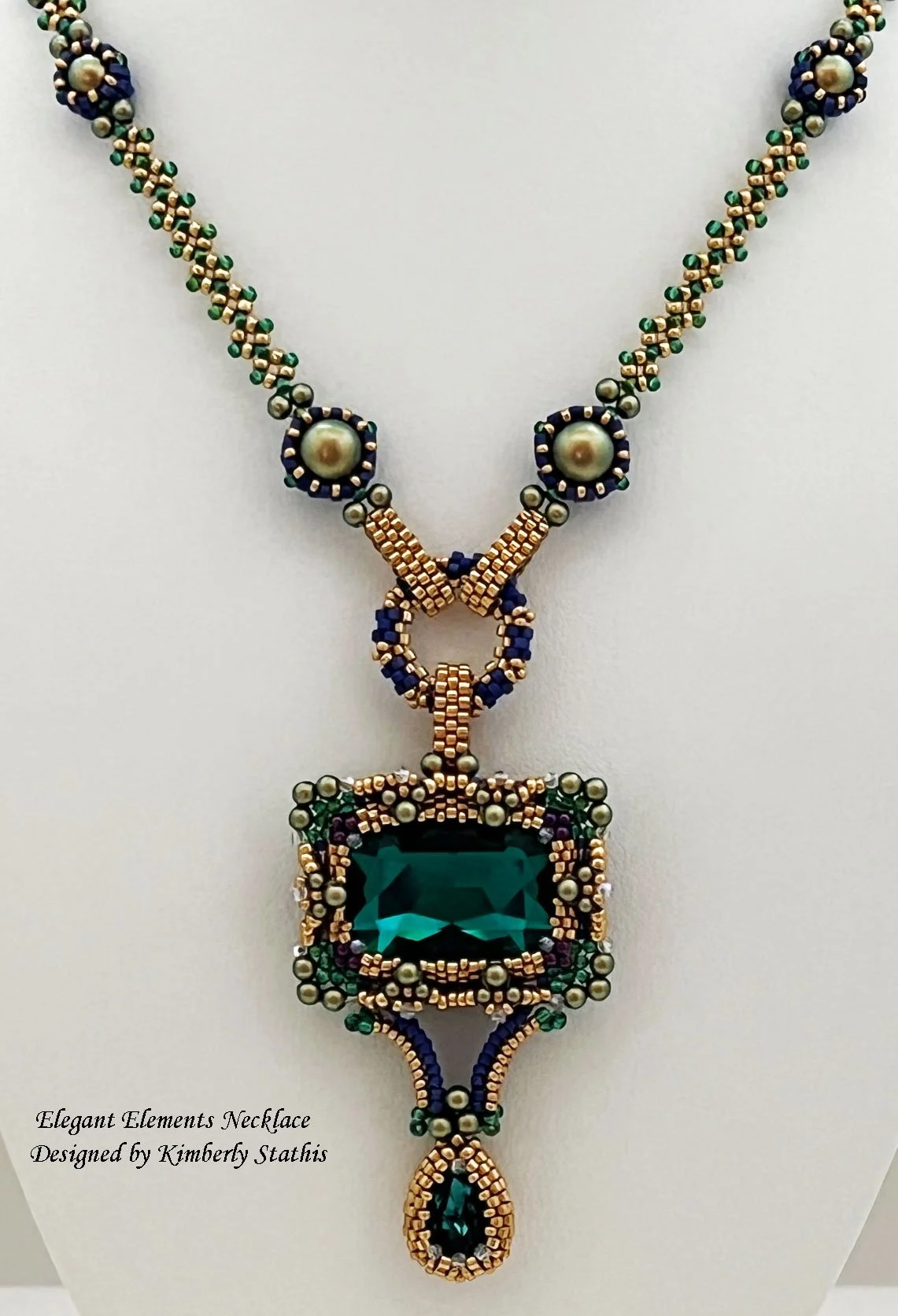 A beaded necklace featuring a large rectangular green gemstone pendant with intricate beadwork in gold, purple, green, and blue. The necklace has a symmetrical design with round pearl-like beads and smaller colorful beads, and a dangling teardrop-sha