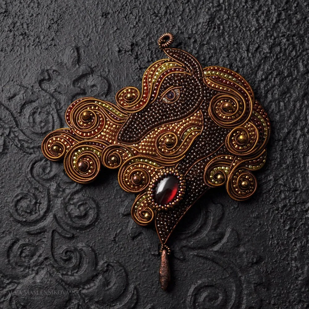 Intricate beaded brooch in the shape of a peacock with swirling feathers, featuring a large red gemstone on a dark textured background.