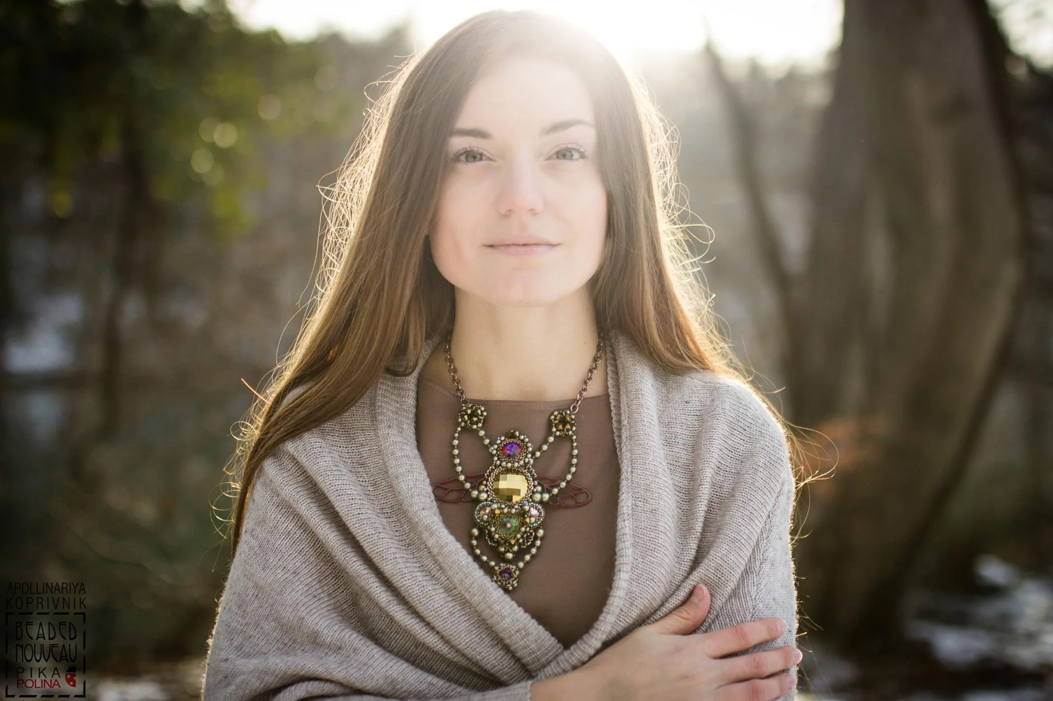 A young woman with long brown hair and light skin is standing outdoors in a natural setting with trees and sunlight. She is wearing a beige sweater and a large, elaborate necklace with colorful stones. She has a gentle smile and is looking at the cam