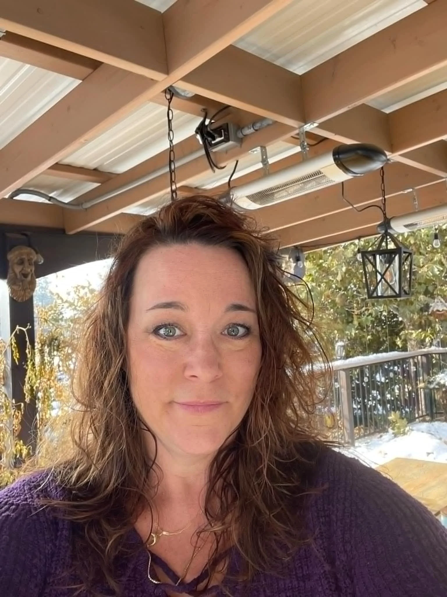 A woman with curly hair and blue eyes taking a selfie outdoors on a porch with wooden beams, decorative lantern, and trees in the background.