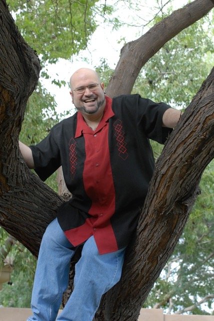 A man with glasses and a bald head smiling, sitting on a tree branch surrounded by green leaves.