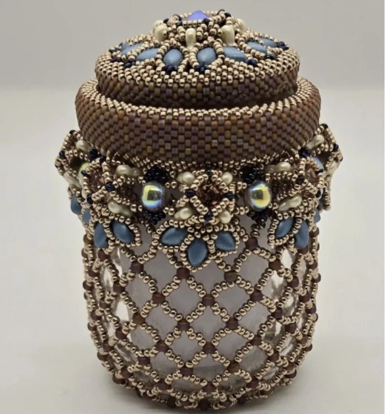 Decorative beaded container with a lid, adorned with intricate beadwork in shades of brown, blue, black, and white, featuring pearl-like beads and floral patterns.