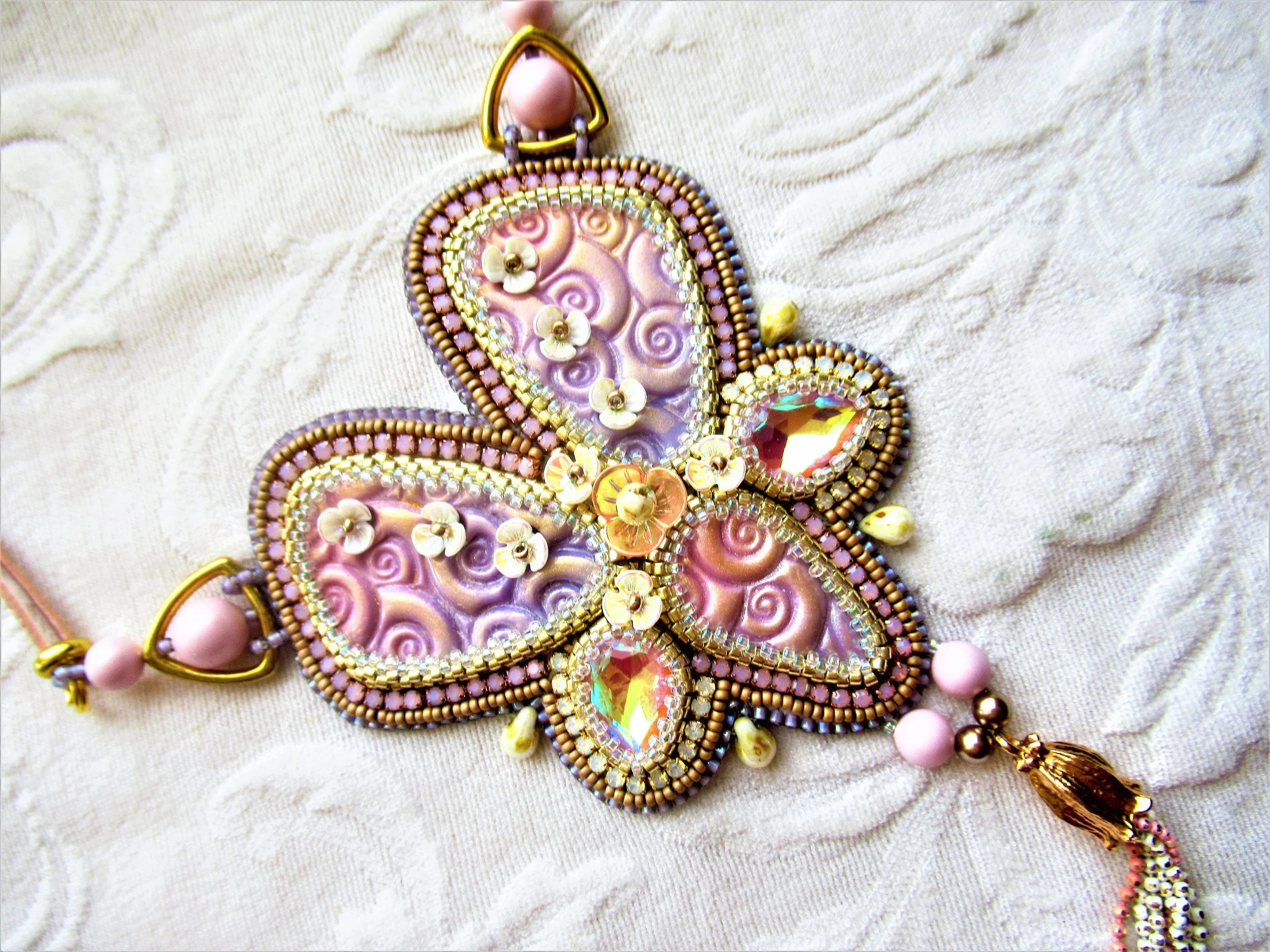 A decorative butterfly-shaped pendant embellished with beads, sequins, pearls, and a large iridescent gemstone, hanging against a fabric background.